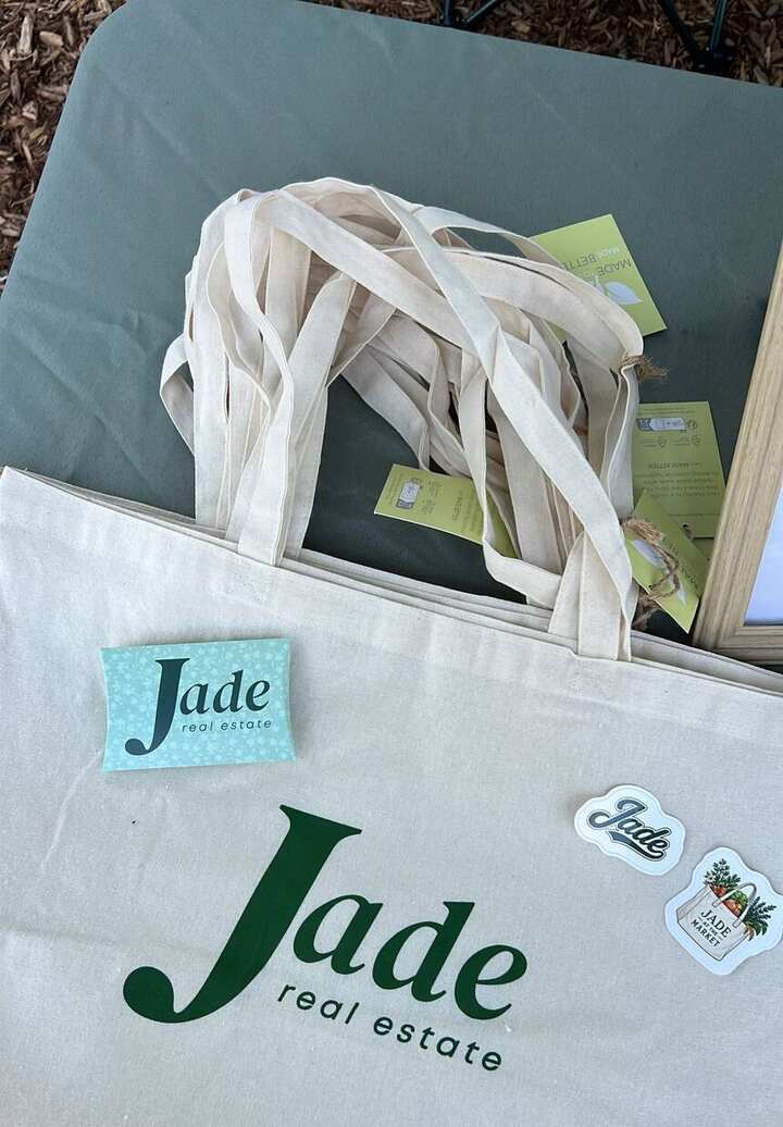 A Jade Real Estate canvas tote bag and branded stickers, including a Jade at the Market sticker, on a market table.